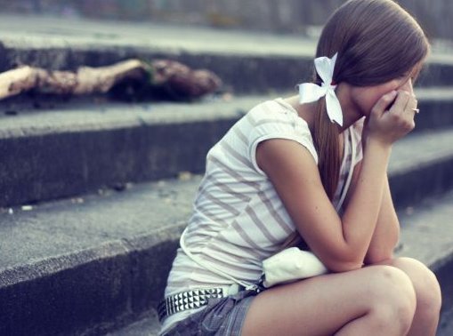 Teenager-Mädchen mit langen braunen Haaren sitzt auf Steintreppe. Sie hat das Gesicht auf die Hände gestützt und sieht traurig aus.